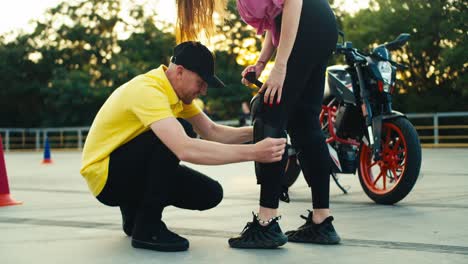 Instructor-in-a-driving-school-in-a-yellow-t-shirt-helps-a-girl-put-on-protective-plates-for-riding-a-motorcycle.-Knee-pads-and-other-protection-for-riding-a-motorcycle