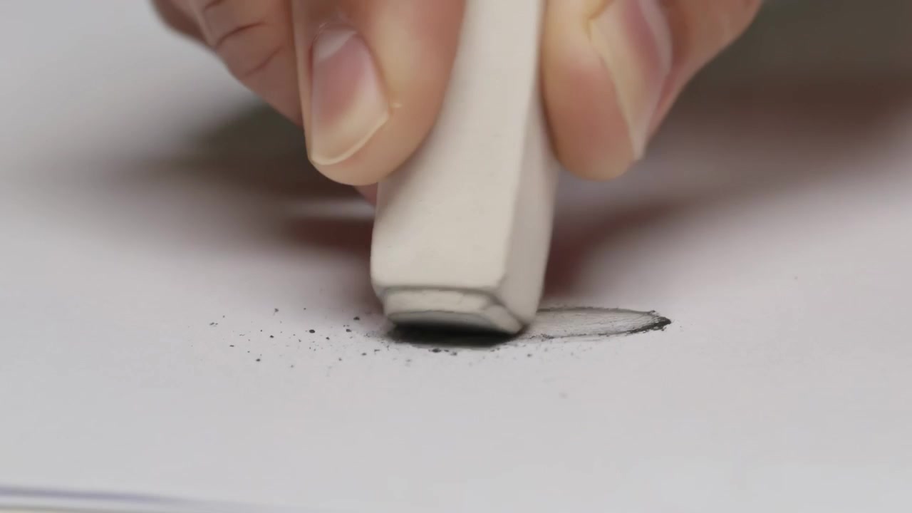 Bajando la mano derecha, guiando una goma de borrar blanca sobre un papel en la mesa, eliminando una marca de grafito y migas.