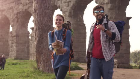 young couple backpackers tourists walking with map guide near roman aqueduct arches in parco degli acquedotti park ruins in rome at sunrise with guitar and sleeping bag slow motion steadycam