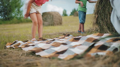 close up of people spreading checkered blanket in grassy field while little boy pulls hay bale in background, rural outdoor picnic preparation under warm sunlight with dry grass