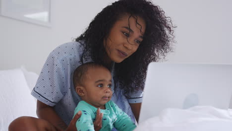 Mother-Wearing-Pyjamas-Cuddling-Baby-Son-In-Bedroom-Whilst-Working-From-Home-On-Laptop