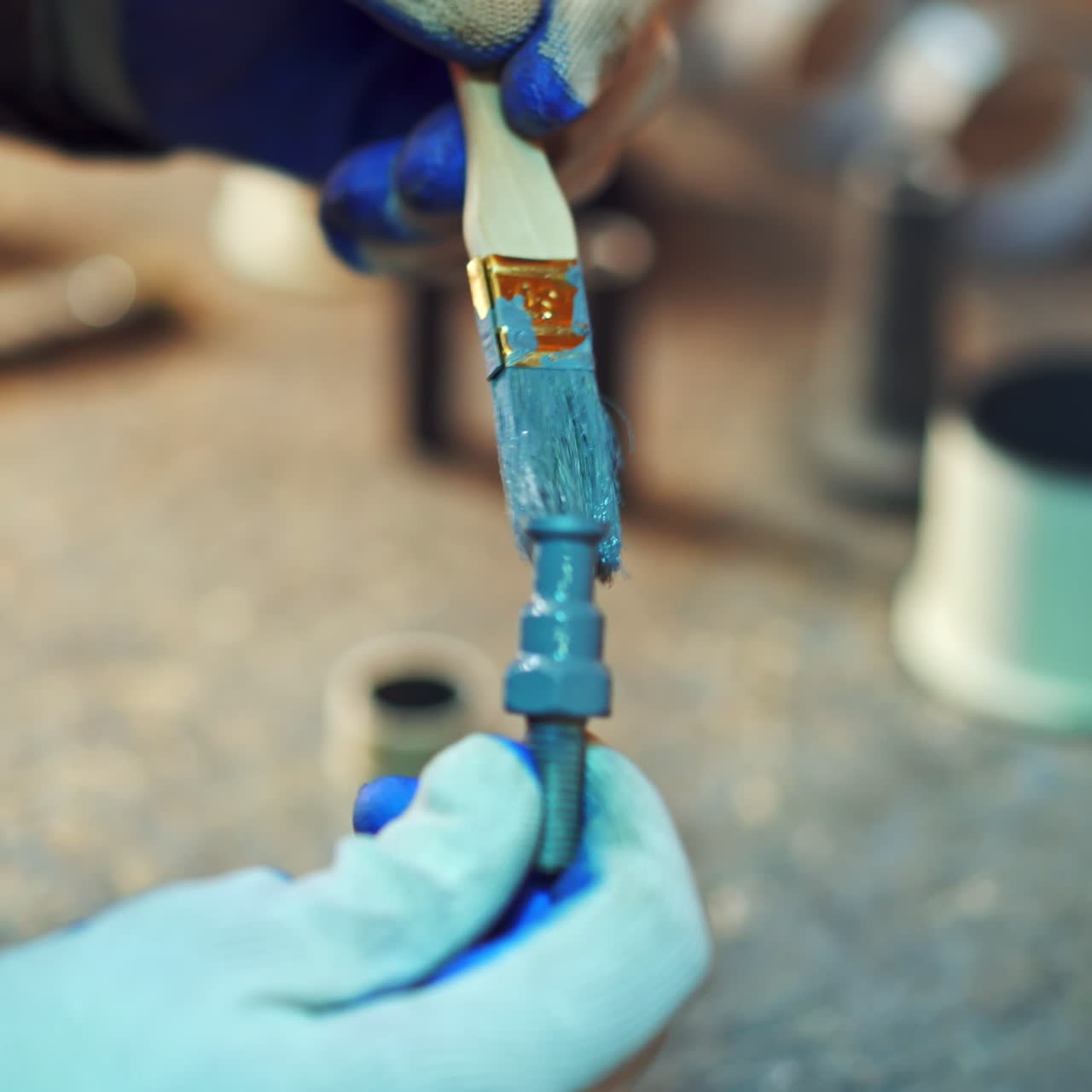 Close-up of man's hands with a brush painting metal tool with blue color in factory. Male worker in gloves paints instruments at workshop.