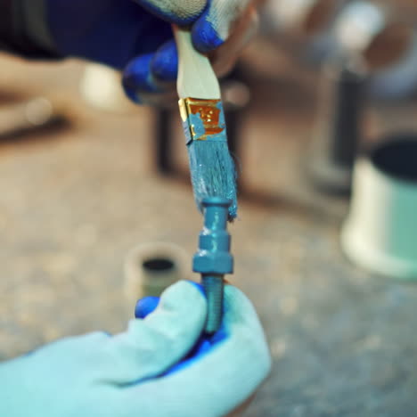 close-up of man's hands with a brush painting metal tool with blue color in factory. male worker in gloves paints instruments at workshop.