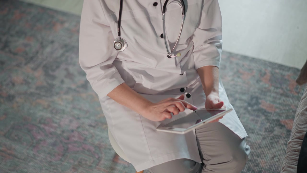 Dokter Wanita Duduk Mencatat di Tablet Saat Kunjungan Perawatan di Rumah, Menulis Rekam Medis Digital Sambil Berinteraksi dengan Pasien. Pemeriksaan Lanjutan Penuh Perhatian di Ruang Tamu Berlantai Karpet.