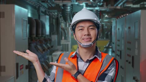 close up of asian male engineer with safety helmet smiling and pointing to side while standing in engine control room, work of electrical generators
