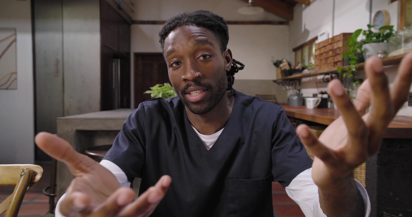 Uomo africano adulto in divisa medica blu scuro che guarda la telecamera al tavolo della cucina, mentre si china e si controlla le mani