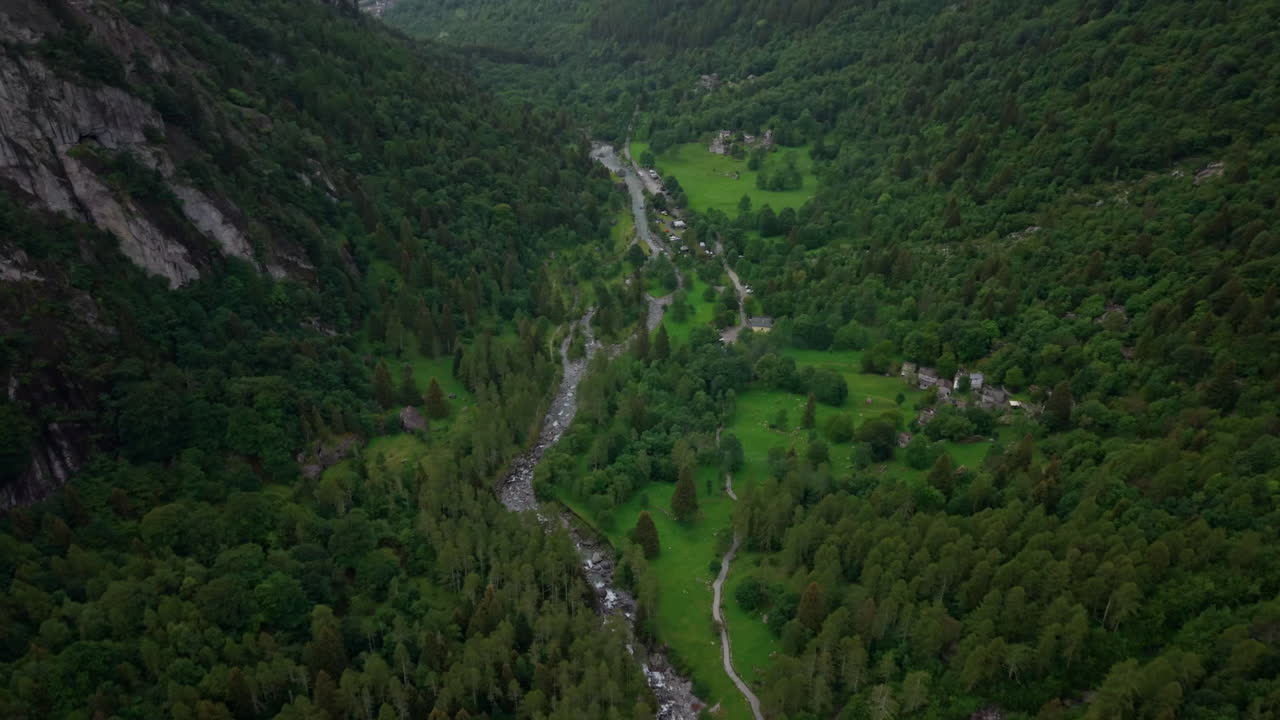 Drone flies forward over a lush alpine valley between two mountains, showing a winding river, scattered houses, and dense forest