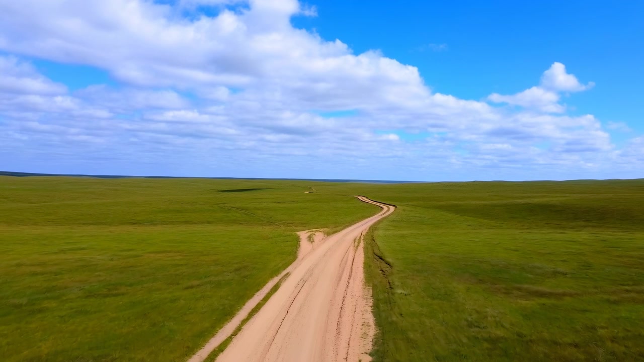 an aerial view of a dirt road leading into the distance, surrounded by a serene open field with lush green grass and brown dirt. the tranquil scene is set against a backdrop of a clear blue.