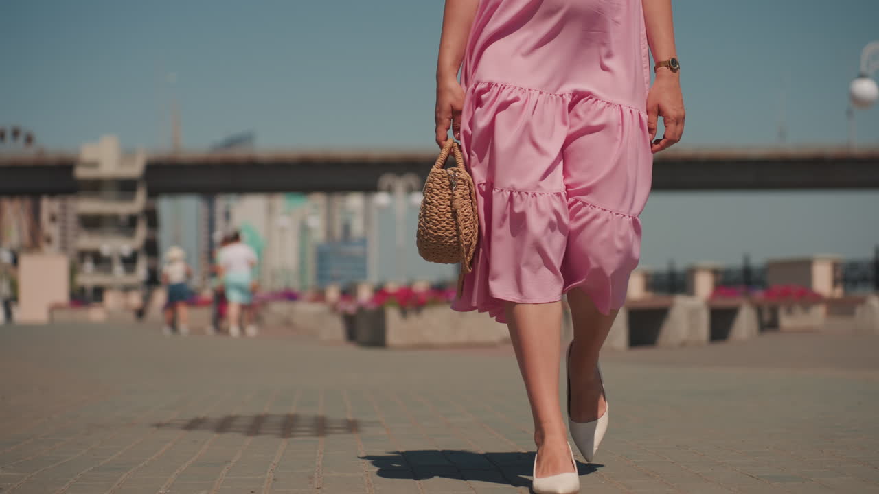 Corpo intero di una donna che cammina sul lungomare, abito rosa svolazzante che ondeggia a ogni passo, piazza urbana soleggiata con ponte e skyline sullo sfondo, sandali casual, sguardo fisso e postura sicura, tempo libero