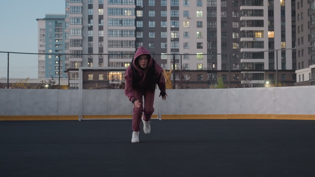 Female athlete on white court line running across black asphalt sports court near white barrier topped with chain link fence in urban setting wearing maroon hoodie and white sneakers
