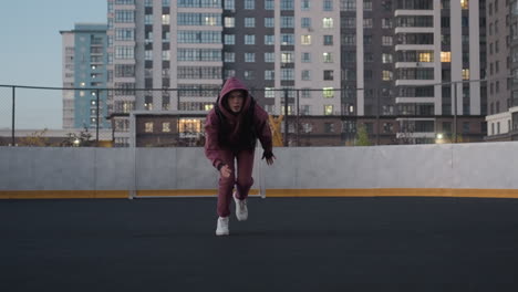 female athlete on white court line running across black asphalt sports court near white barrier topped with chain link fence in urban setting wearing maroon hoodie and white sneakers