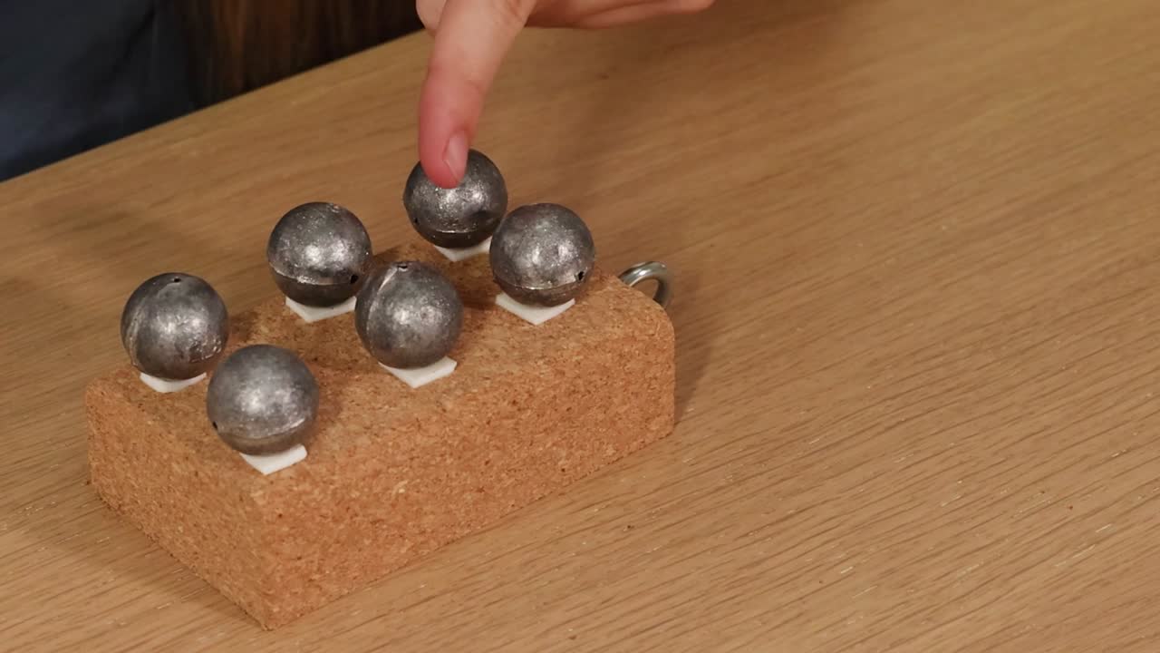 A person's hands interact with several small metal balls arranged on a textured cork block