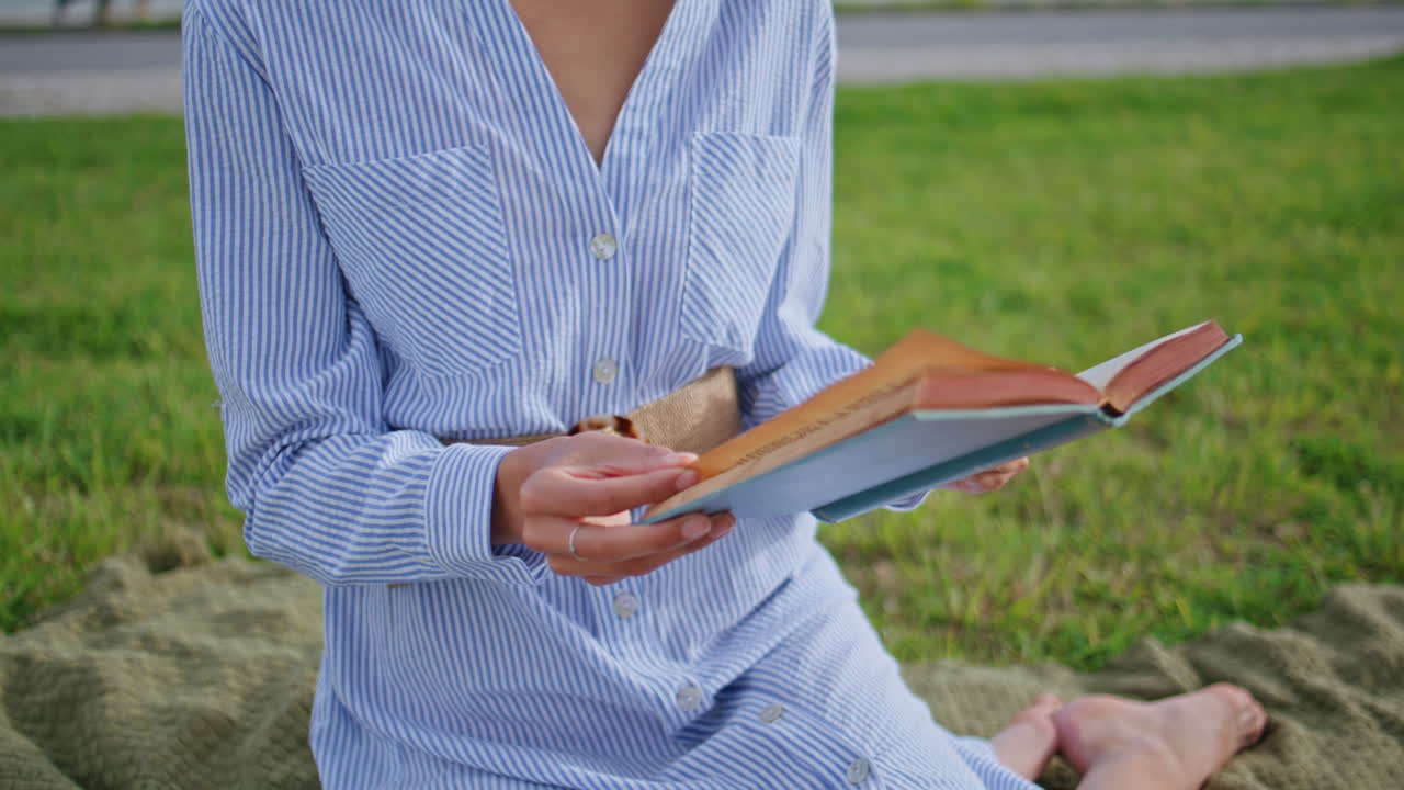 Closeup lady reading book on picnic blanket surrounded by greenery. Relaxed girl