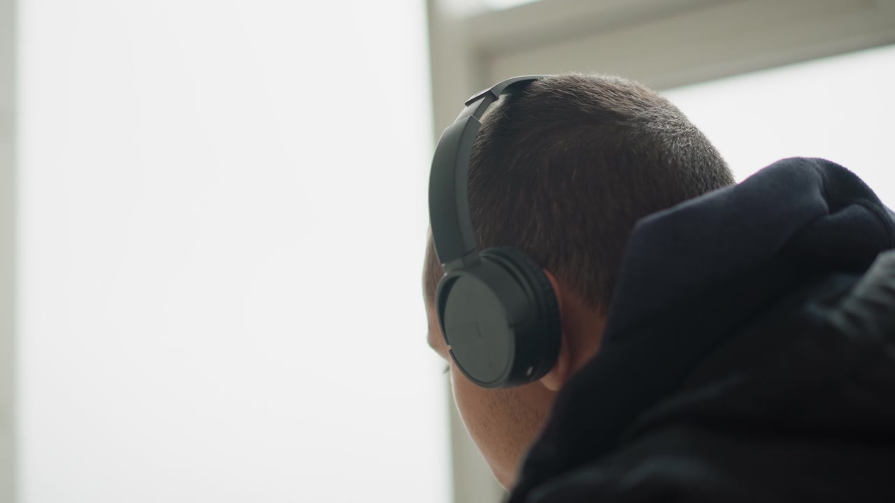 Nærbilde av mann iført svarte hodetelefoner og mørk hettegenser, fokusert på skjermen med mykt naturlig lys fra et vindu. Hodetelefonene er av over-øret-typen, og personen holder en jevn posisjon.