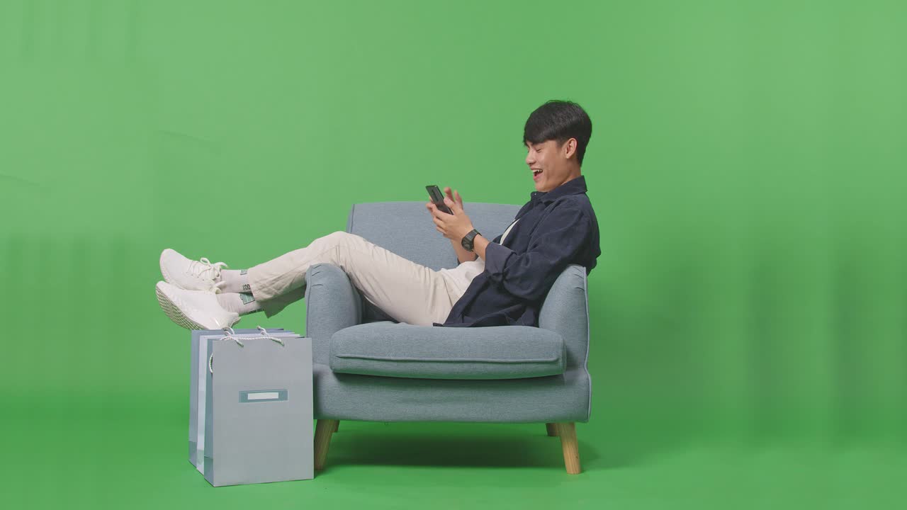 쇼핑 가방 을 들고 있는 아시아인 쇼핑맨 이 녹색 스크린 앞 에 소파 에 앉아 휴대 전화 로 온라인 쇼핑 을 즐긴다