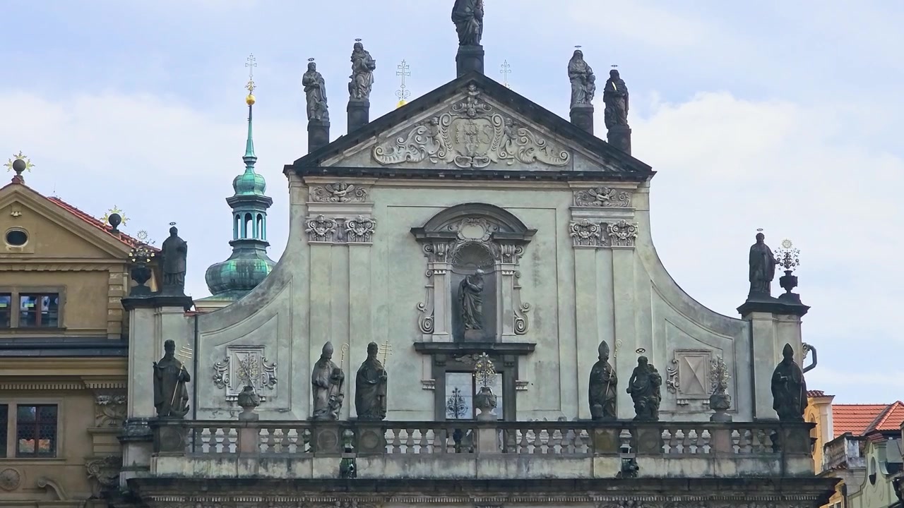 Widok z przodu zabytkowej barokowej fasady kościoła w Pradze, Czechy, z kamiennymi posągami, ozdobnymi rzeźbami religijnymi, łukowatą architekturą, dachami starego miasta i delikatnym błękitnym niebem.
