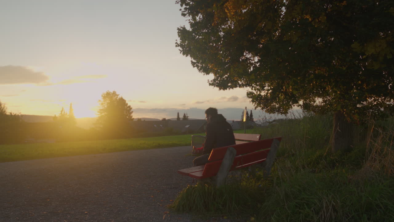 Young man with a backpack sits down on a bench to enjoy the sunset view, Switzerland