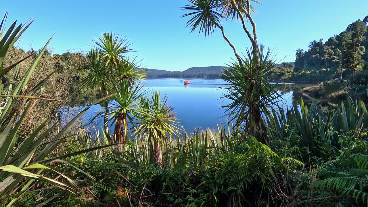 View of Lake Mapourika in New Zealand through greenery as a boat sails toward a calm bay. The clear water reflects the summer sky on a peaceful day