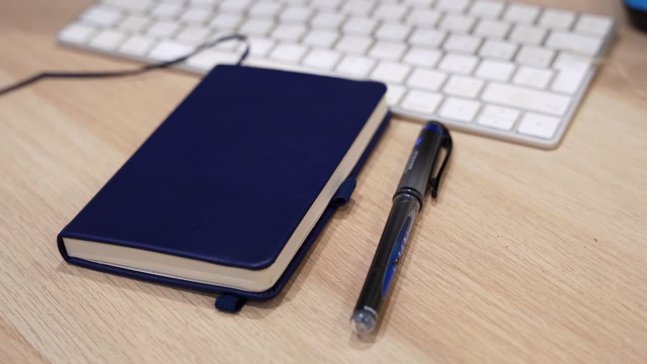 Buku catatan biru dengan pena hitam di samping keyboard komputer di meja kayu di dalam ruangan, menampilkan fokus dangkal dan komposisi ruang kerja yang bersih