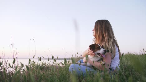 young happy woman and het little dog sitting with flying kite on a glade at sunset