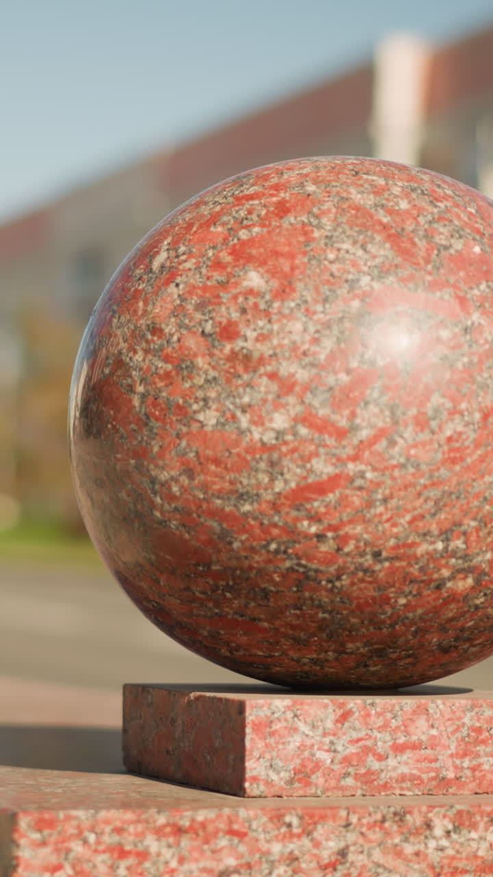 햇빛이 비치는 받침대 위의 붉은 화강암 구, 도시 광장의 클로즈업은 반짝이는 얼룩덜룩한 표면을 보여주고 따뜻한 햇빛이 비치며, 흐릿한 건물 배경, 얕은 피사계 심도, 석재 블록 받침대, 반사