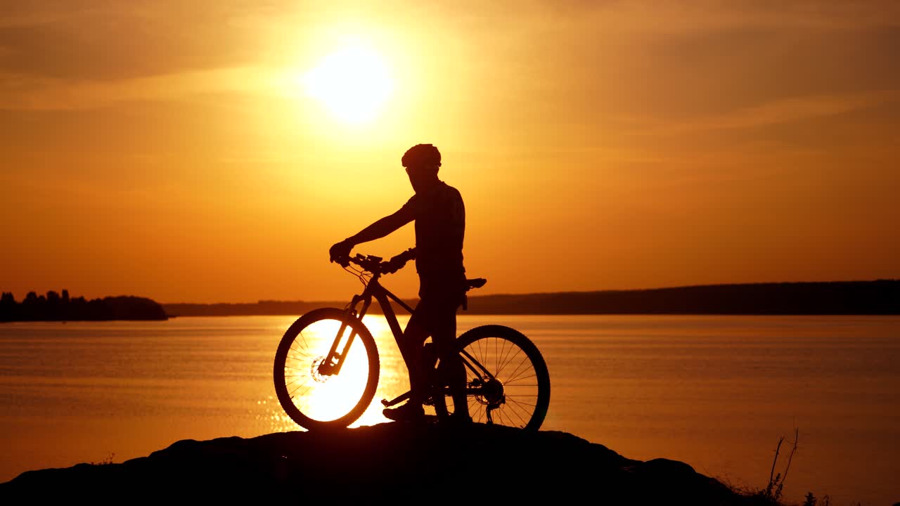 Silhouette of a male cyclist with helmet at sunset near the river. Bike rider walks with his bike on the background of the golden path from the sun over the water surface.