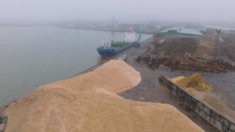 aerial establishing view of wood terminal crane loading timber into the cargo ship, port of liepaja , lumber log export, overcast day with fog and mist, wood chip piles, wide drone moving back