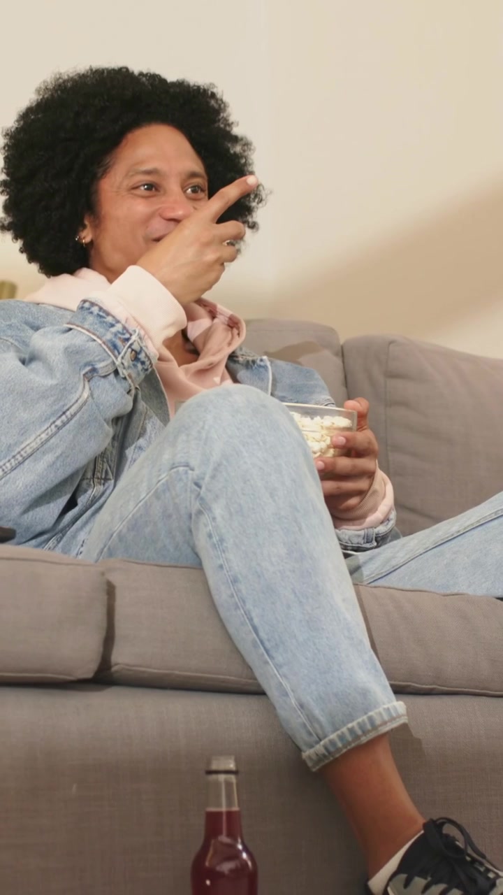 Video verticale: Uomo che guarda un film d'azione in TV, punta il telecomando, ride e mangia popcorn in jeans sul divano