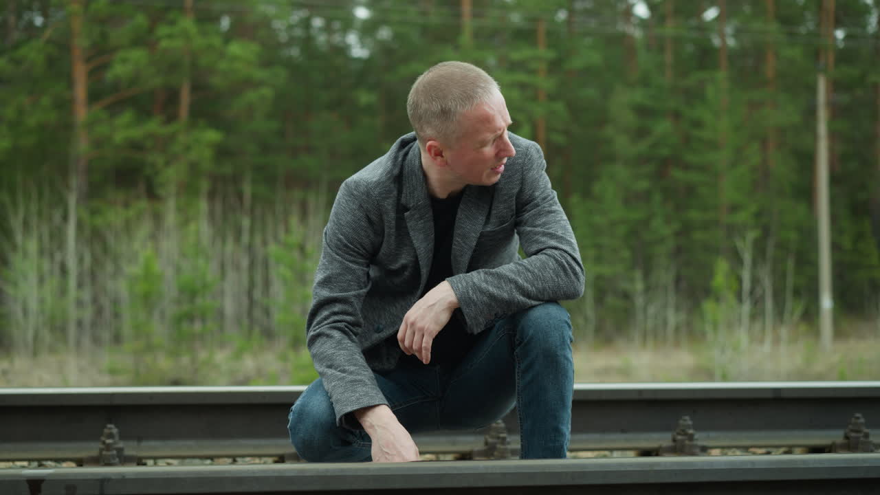 A close-up view of a man in a grey blazer and blue jeans kneeling by a railway track, examining it closely with a thoughtful expression