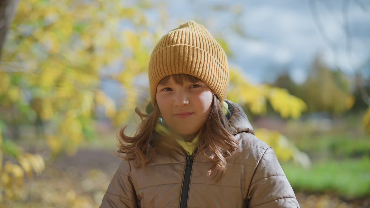 cute child wearing warm hoodie and mustard beanie standing outdoors with soft smile, surrounded by autumn scenery and golden leaves gently swaying in wind under bright sky