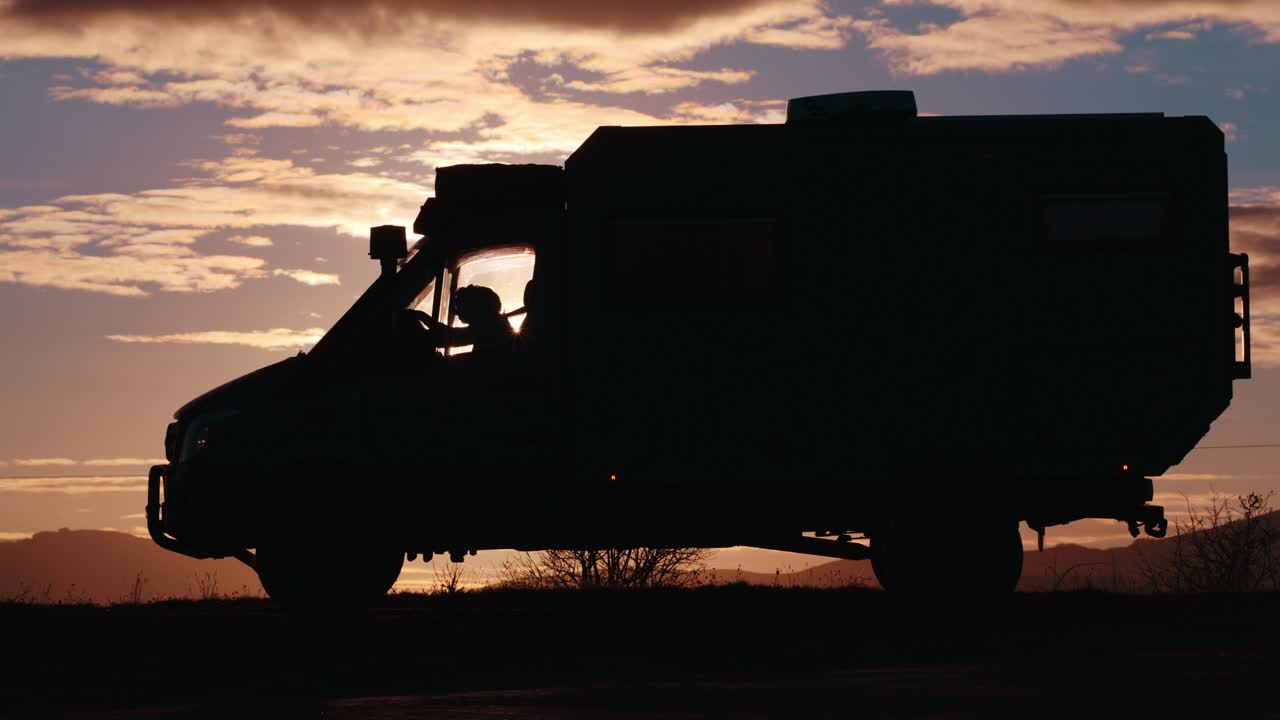 An expedition truck silhouetted against a dramatic golden hour sunset. A driver is visible inside, parked on a mountain, evoking adventure and van life