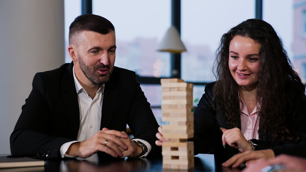 Curly-haired woman pulls a little brick from tower in jenga game. Friends sitting next to her try to distract and make her fail.
