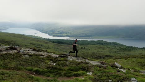 Vista-Aérea-De-Seguimiento-De-Un-Hombre-Corriendo-A-Lo-Largo-De-La-Cresta-De-La-Colina-De-Ullapool-Con-El-Lago-Achall-Al-Fondo