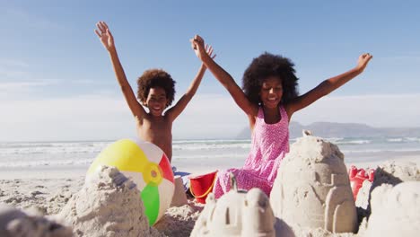 sonrientes hermano y hermana afroamericanos con los brazos en el aire, jugando con arena en la playa