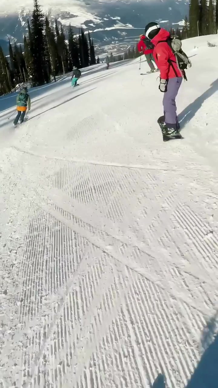 Video verticale: Snowboarder in giacca rossa che fa carving su una pista battuta, la telecamera segue gli amici.
