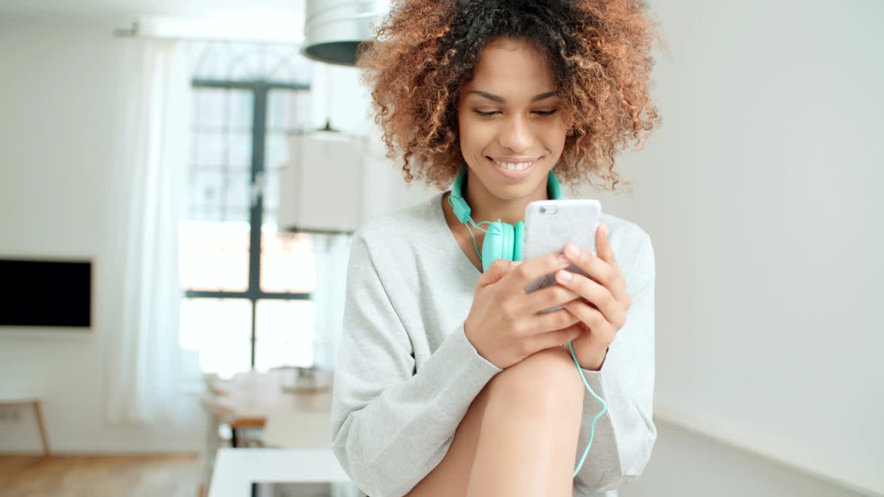 una hermosa joven afroamericana enviando mensajes de texto por teléfono móvil en casa.