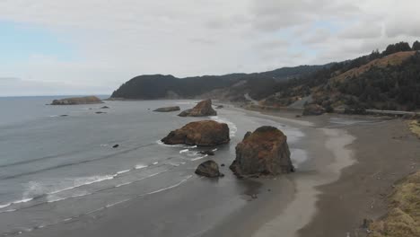 Dreamy-aerial-of-Oregon-coastline-with-large-rock-outcroppings-and-waves-lapping-up-on-a-natural-beach