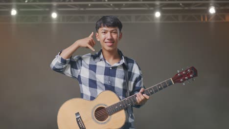 adolescente asiático con guitarra mostrando me llama gesto y sonriendo en el escenario