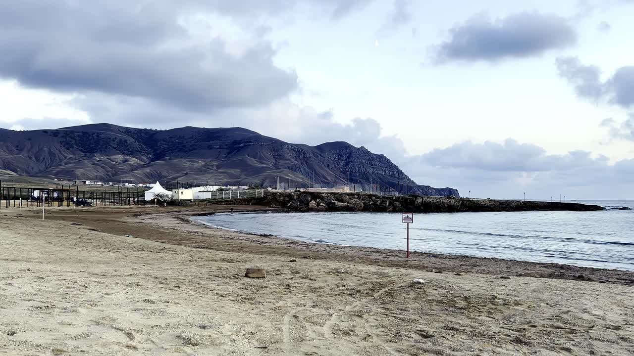 해가 지는 순간에 평온한 해변이 펼쳐져 있고, 바가에 부드러운 파도가 부히고, 먼 곳에서 구름이 가득한 하늘 아래 바위 산이 아져서 조용한 분위기를 조성합니다.