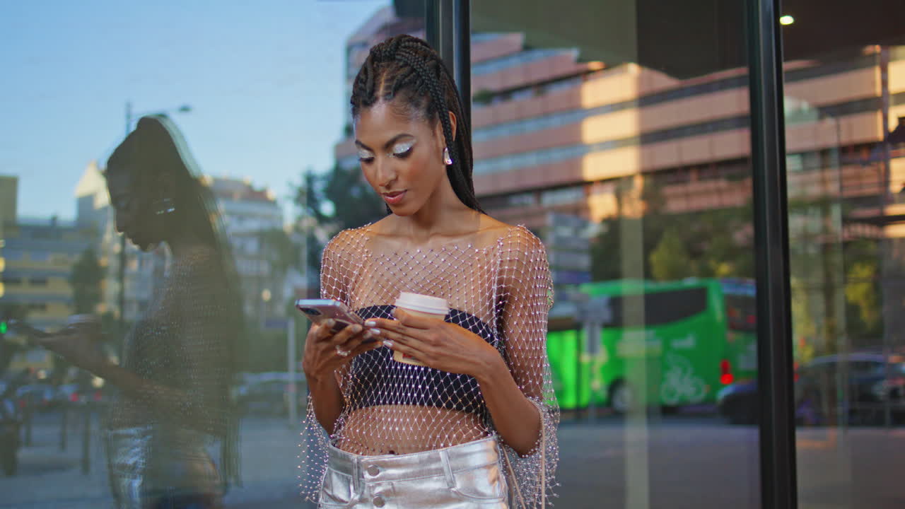 Primo piano di modella dalla pelle scura che digita sullo smartphone vicino a una parete di vetro. Donna che si fa un selfie.