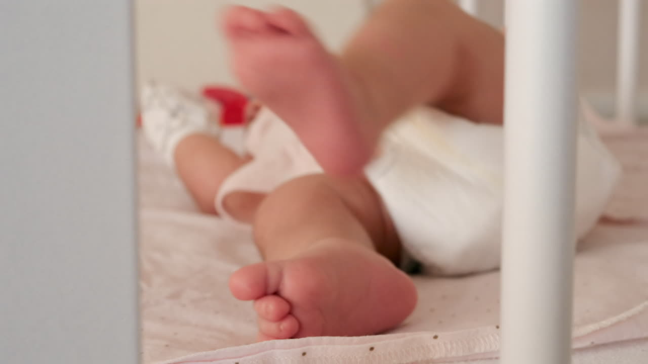 Close up of a baby's legs and diaper as the infant rests in a crib, seen through the crib bars