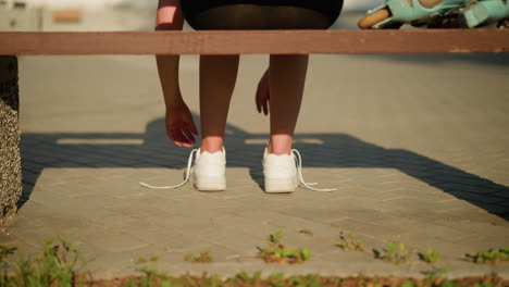 back leg view of lady loosening shoelaces of white sneakers while seated on bench under bright sunlight, with soft shadows cast on pavement and roller skate partially visible on bench