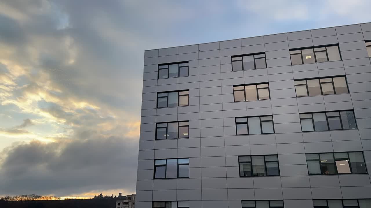 Video timelapse di un moderno edificio aziendale grigio con finestre a griglia durante il tramonto, nuvole in rapido movimento che attraversano un cielo spettacolare e luce dorata all'orizzonte in un contesto urbano