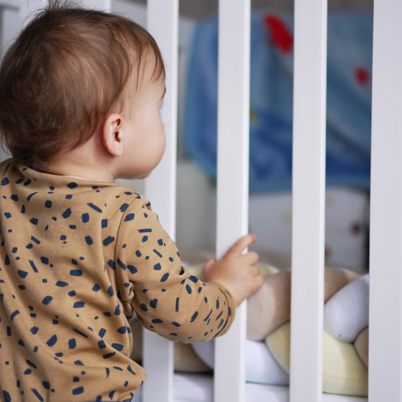 Nice little Caucasian baby stands holding by his white crib. Adorable kid looks backwards into camera
