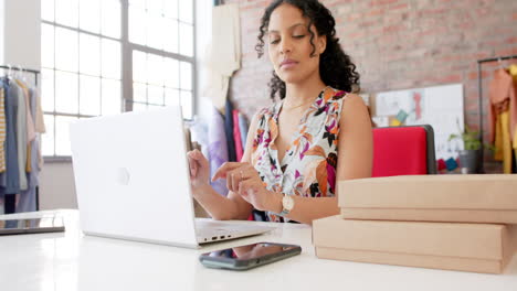 diseñadora de moda bi-racial feliz usando portátil y teléfono inteligente en el estudio, cámara lenta