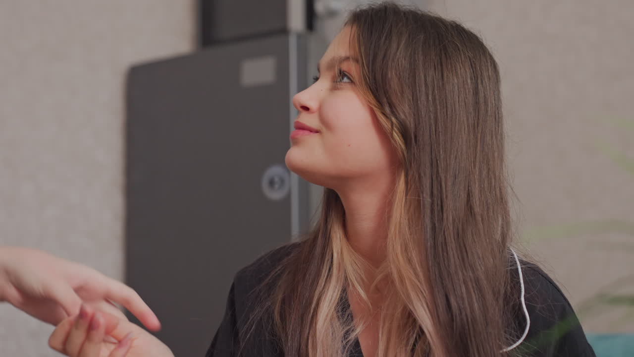 girl with long dark hair sits near green decorative plant holding sliced cheese, chewing and looking away while interacting with someone out of frame in cozy modern kitchen with soft lighting
