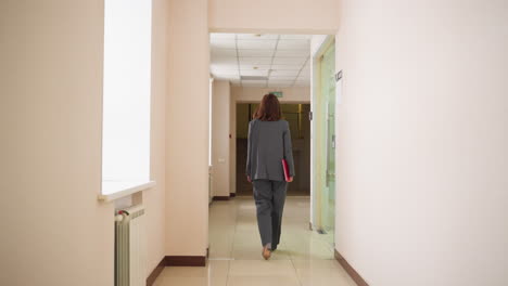 Businesswoman in gray suit walking down office hallway holding red folder heading to meeting or presentation with focused posture in modern corporate workspace during professional workday