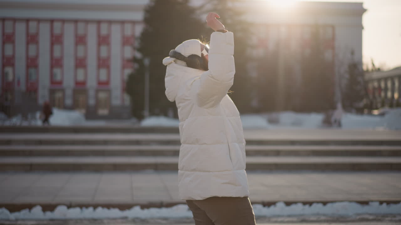 Side view of elegant young woman overflowing with excitement as she sways joyfully on sunlit winter pavement wearing puffy coat and headphones while rays of setting sun glow behind her movement