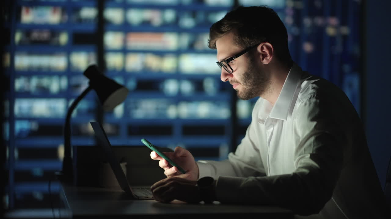 Office Building with Businessman Using Mobile Phone and Standing by the Office Window. Man working in modern office late at night. Busy caucasian manager at work with mobile telephone.