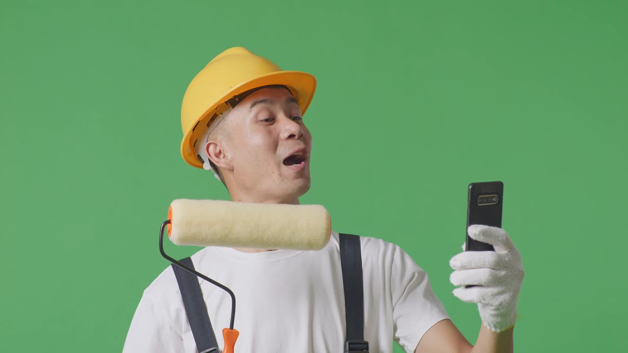 Close Up Of Asian Man Painter Wearing Safety Helmet Waving Hand Having A Video Call On Smartphone While Standing In The Green Screen Background Studio